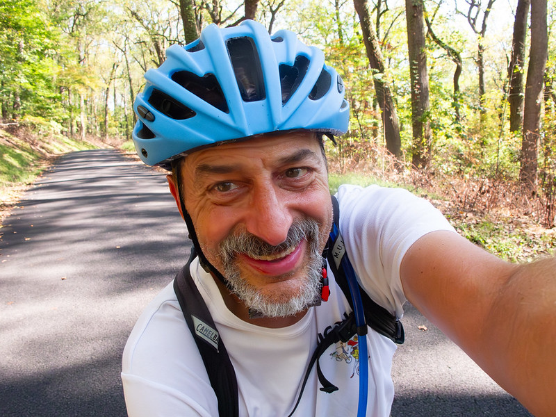 A smiling man in a bicycle helmet holds out the camera ahead of him for a selfie. The road behind curves through a bright forest, quiet and dotted with fallen leaves.