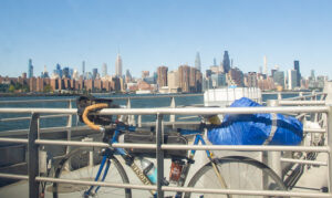 A picture shot through the window of a ferry. A Marinoni bicycle with touring luggage is leaning against a railing at the front of the boat. The skyline of New York City is seen in the background, with the Empire State Building in the middle