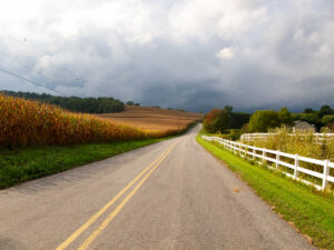 A rural road winds around hills, with a corn field on the left and a white picket fence on the right. The sky is unsettled, with a possible storm on the horizon.